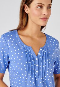 Top corto de manga corta azul con lunares blancos, que presenta un cuello henley con cierre de botones y sutiles detalles plisados. Textura de tela suave.