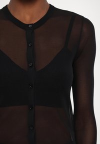 Gros plan d'une personne portant un haut noir transparent à manches longues boutonné sur un caraco noir à fines bretelles.