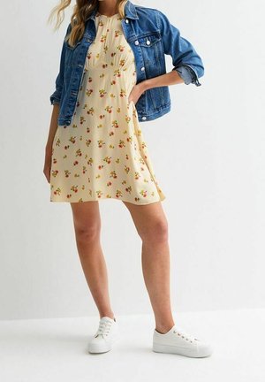 Kobieta w żółtej kwiecistej sukience do kolan, niebieskiej jeansowej kurtce z podwiniętymi rękawami i białych trampkach, stojąca z jedną ręką na biodrze.