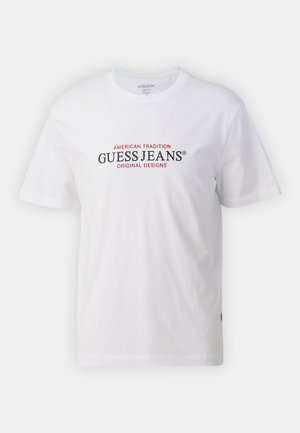 Witte katoenen t-shirt met een ronde hals en korte mouwen, met rode en zwarte tekst die "GUESS JEANS" en "AMERICAN TRADITION" lezen.