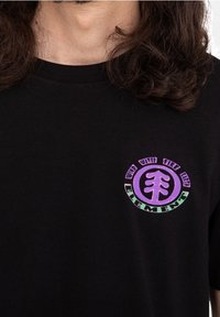 T-shirt en coton noir à manches courtes. Présente un graphisme circulaire en violet et vert avec les mots "VENT EAU FEU TERRE ÉLÉMENT."
