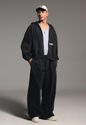 Ung mand iført en hvid kasket, sort hættetrøje med lynlås, grå skjorte, vide sorte bukser og hvide sneakers, stående med hænderne i lommerne.