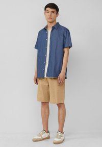 Camicia azzurra in denim a maniche corte, con colletto, abbinata a pantaloni corti beige. T-shirt bianca sotto e sneakers chiare.