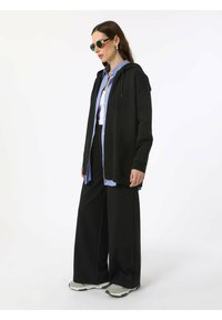 Black zip-up hoodie over a blue striped shirt, paired with wide black pants and grey sneakers; model wearing round sunglasses.