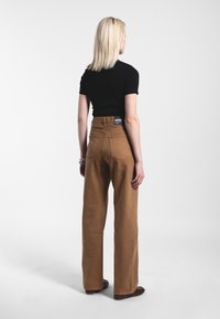 Weite braune Denim-Hosen mit hoher Taille, ausgestattet mit Gesäßtaschen und einem Etikett am Bund. Kombiniert mit einem schwarzen, figurbetonten Oberteil.