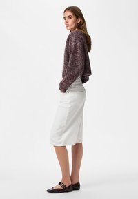 Jeune femme debout de profil, portant un pull court violet, une jupe blanche longueur genou et des chaussures plates foncées sur un fond uni.