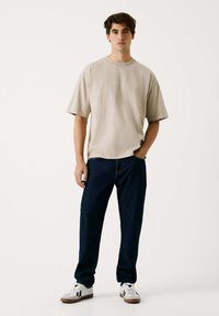 Beige übergroßes T-Shirt mit kurzen Ärmeln, Rundhalsausschnitt und subtilen Textdetails, kombiniert mit dunkelblauen Jeans und weißen Sneakers.