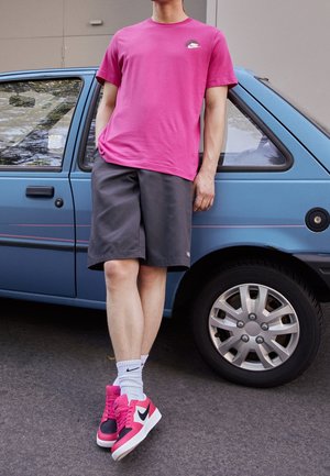 Osoba opierająca się o niebieski samochód, ubrana w różową koszulkę Nike, czarne szorty, białe skarpetki Nike oraz różowo-czarne buty sportowe Nike.