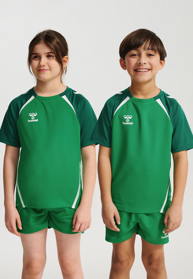 Dwoje uśmiechniętych dzieci stoi obok siebie, ubrane w zielone sportowe koszulki i spodenki z białymi akcentami na jednolitym tle.