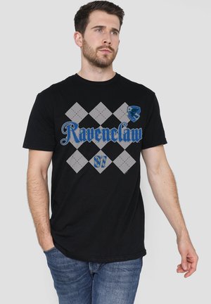 Mann mit kurzen dunklen Haaren und Bart, trägt ein schwarzes Ravenclaw-T-Shirt und blaue Jeans, schaut leicht nach rechts mit der Hand in der Tasche.
