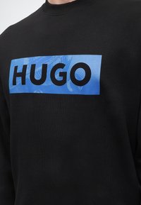 Personne portant un sweat-shirt noir avec un logo rectangulaire bleu affichant le mot « HUGO » en lettres noires grasses sur la poitrine.