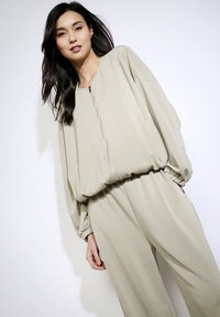 Set di abbigliamento da casa color beige chiaro, composto da una maglia con zip dal taglio morbido, maniche voluminose e polsini elasticizzati, abbinata a pantaloni larghi.