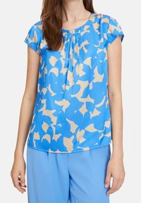 Kvinna klädd i en blå kortärmad blus med beige abstrakta mönster och matchande blå byxor, stående med armarna avslappnat längs sidorna.