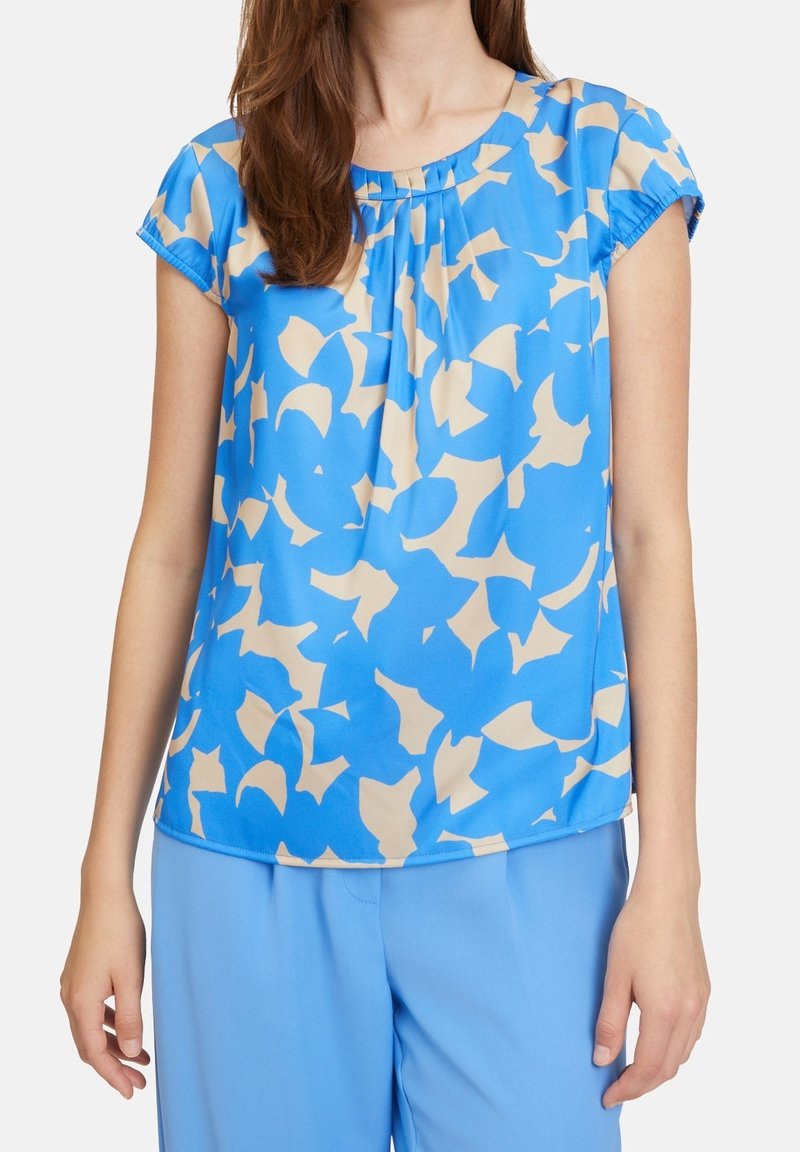 Kvinna klädd i en blå kortärmad blus med beige abstrakta mönster och matchande blå byxor, stående med armarna avslappnat längs sidorna.