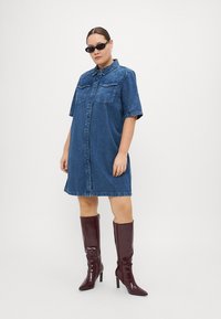 Denim shirtjurk in blauw met korte mouwen, knoopsluiting en twee vo pockets. In combinatie met kniehoge bordeauxrode lakleren laarzen.