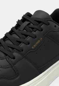 Μαύρο δερμάτινο αθλητικό παπούτσι με επίπεδα μαύρα κορδόνια, λείο φινίρισμα και αντίθετη λευκή λαστιχένια σόλα. Χρυσή λεπτομέρεια λογοτύπου κοντά στους οφθαλμούς.