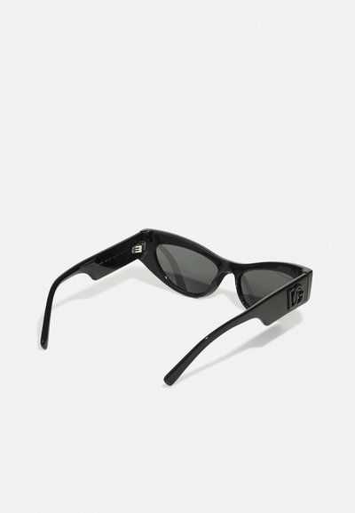 Lunettes de soleil noires au format anguleux de type œil de chat et aux verres sombres. Elles présentent des montures brillantes noires et un logo stylisé sur le côté.