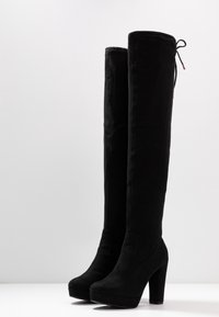 Stivali neri sopra il ginocchio in morbido suede, con tacco alto chunky, suola piattaform e un nastro decorativo in cima.