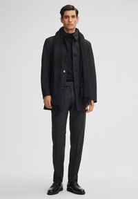Zwarte wollen jas met knopen, gelaagd over een zwarte turtleneck, gecombineerd met donkere op maat gemaakte broek en zwarte schoenen. Sjaal heeft franjes aan de randen.