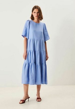 Nainen seisomassa vaaleansinisessä kerroksittaisessa midimekossa, jossa on lyhyet pöyhistetyt hihat ja ruskeat korolliset sandaalit yksiväristä taustaa vasten.