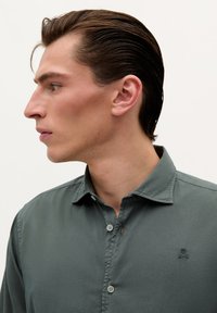 Camisa de hombre de botones en verde apagado. Cuenta con collar, dos botones, un sutil logo bordado en el pecho y una tela suave, ligeramente texturizada.