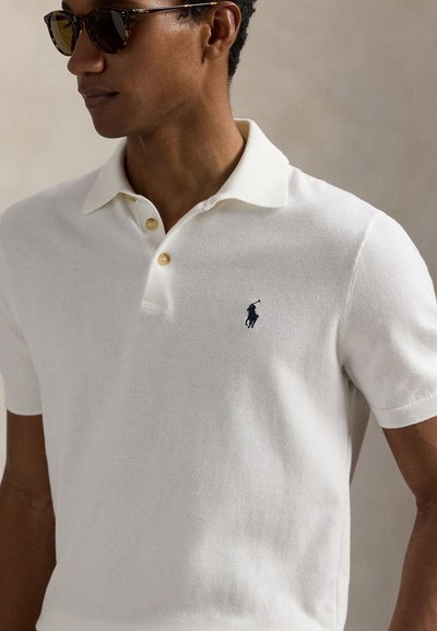 Baltas polo marškinėliai iš minkštos mezgimo medžiagos, su klasikine apykakle, dviem mygtukais ir tamsiai mėlynu siuvinėtu logotipu ant krūtinės.