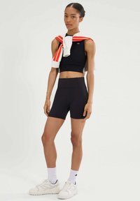 Zwarte sportieve crop top en hoge taille shorts, gecombineerd met witte sneakers en crew sokken. Een wit en rood sweater ligt over de schouders.