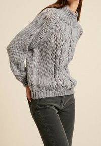 Femme portant un pull en maille épaisse gris clair avec des motifs torsadés et un pantalon en velours côtelé gris foncé, sur un fond neutre.