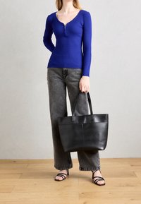 Haut bleu à manches longues côtelé avec une fermeture éclair, jean gris à jambes larges, et sac fourre-tout en cuir noir avec deux poignées courtes. Fond en bois.