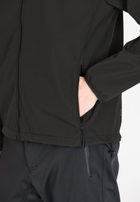 Musta pehmeäkuorinen takki, jossa on vetoketjullinen etuosa, sivutaskut ja säädettävät hihansuut. Ominaisuuksiin kuuluu sileä pinta ja vartalonmyötäinen muotoilu.