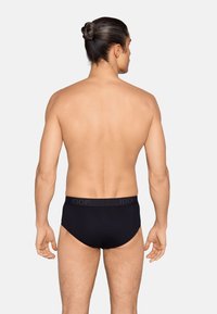 Homme aux cheveux attachés vu de dos portant un slip noir de la marque Joop! sur un fond clair uni.