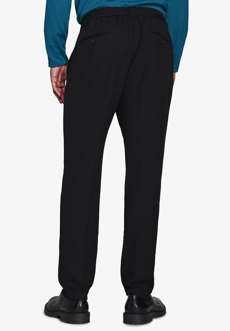 Sorte bukser med tapered fit, der har en stræk-talje og to baglommer. Båret med sorte sko på en hvid baggrund.