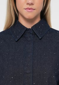 Tumman sininen denimmshirt klassisella kauluksella ja hienovaraisilla hopeapilkkuilla. Erottuva etuvetoketju ja vastakkain ommeltuja saumoja.