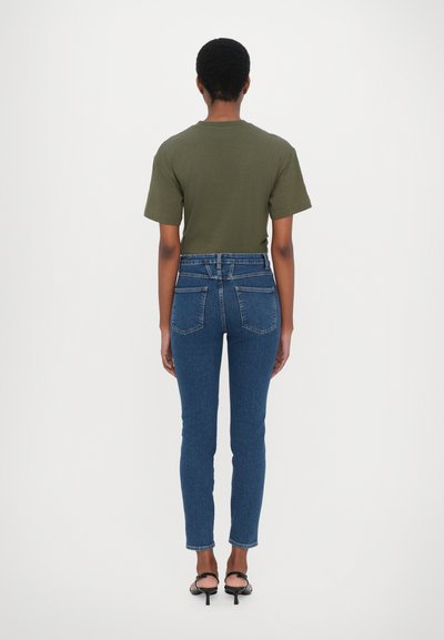 Olivově zelené tričko s krátkým rukávem spárované s tmavě modrými kalhotami s vysokým pasem a zúženým střihem. Outfit má jednoduchý design a hladkou texturu.