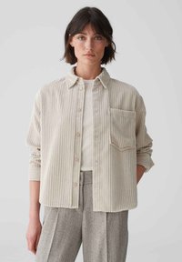 Corduroy button-up shirt in light beige with vertical stripes, chest pocket, and rolled sleeves, layered over a white ribbed shirt.