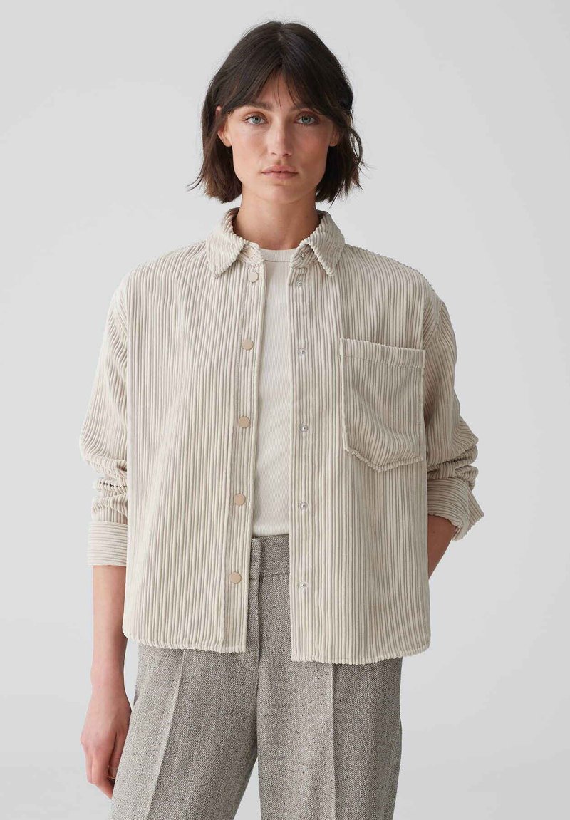Corduroy button-up shirt in light beige with vertical stripes, chest pocket, and rolled sleeves, layered over a white ribbed shirt.