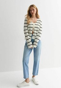 Strickjacke mit schwarzen Streifen auf einem cremefarbenen Hintergrund, mit einer verkürzten Silhouette, kombiniert mit hellblauen Jeans und weißen Turnschuhen.