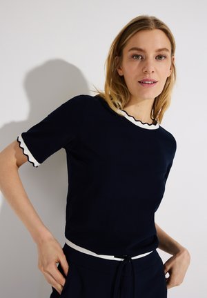 Blondynka ubrana w granatowy krótki rękaw top z białymi falistymi krawędziami i pasujące granatowe spodnie na tle jednolitej białej ściany.