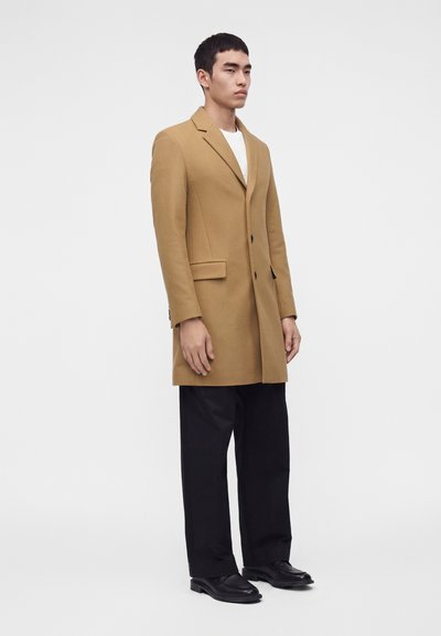 Homme debout portant un long manteau couleur camel, un pantalon noir, des chaussures noires et une chemise blanche sur un fond clair uni.