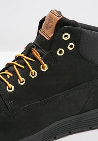 Bottine en daim noir avec lacets jaunes et rouges, œillets dorés et écusson logo en cuir marron. Semelle en caoutchouc texturée et robuste.