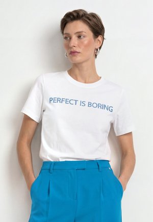 Žena s krátkymi vlasmi nosiaca strieborné kruhové náušnice, biele tričko s textom "PERFECT IS BORING" a jasne modré nohavice s vysokým pásom.