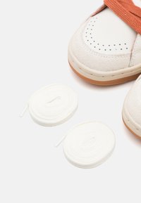 Hvite og beige joggesko med semsket tekstur, oransje stropper og perforert detaljer. Inkluderer to hvite lisser ordnet i sirkler.