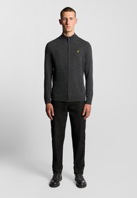 Grijze zip-up trui met hoge kraag en een klein geel logo op de borst. Draagt bij zwarte broek en stevige zwarte schoenen.