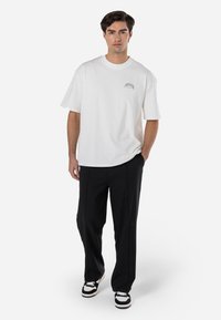 White cotton t-shirt with a crew neck and short sleeves, featuring a small black graphic on the chest. Paired with loose-fitting black trousers.