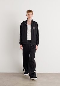 Zwarte sportieve trackjack en broek met witte zijstrepen en logo, gedragen over een witte tanktop, gecombineerd met zwarte schoenen.