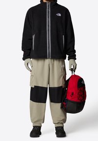 Svart fleecejacka med dragkedja, beige och svarta cargo-byxor, grå handskar, svarta stövlar och en röd ryggsäck med svarta detaljer.