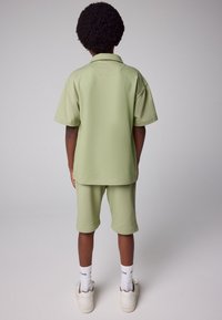 Bagfra billede af et barn iført en lysegrøn kortærmet polo og matchende shorts, kombineret med hvide sokker og beige sneakers.