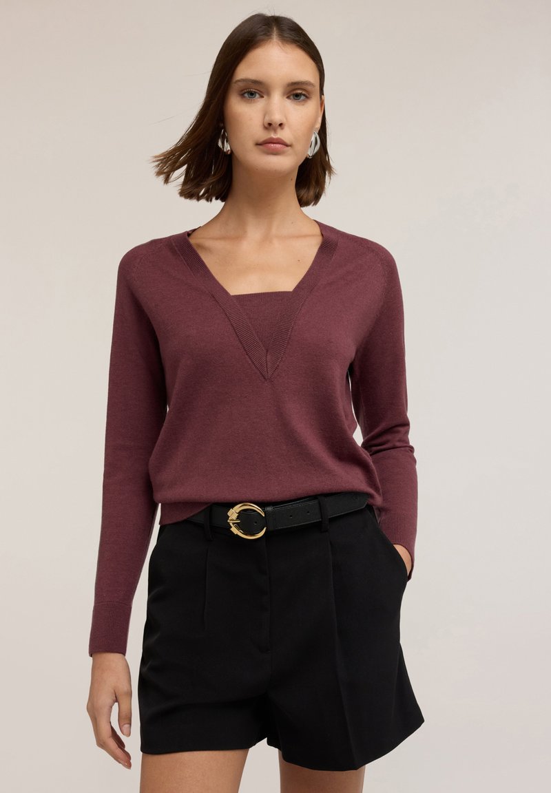 Pitkähihaista viininpunaista villapaitaa, jossa syvä V-pääntie, on kerrostettu neliöpääntiepaidalle. Asusteena on korkeavyötäröiset mustat shortsit ja kultainen vyönsolki.