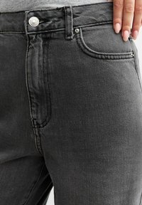 Detailní záběr osoby v šedých denimových džínách s knoflíkovým pasem, ruka odpočívající blízko přední kapsy.
