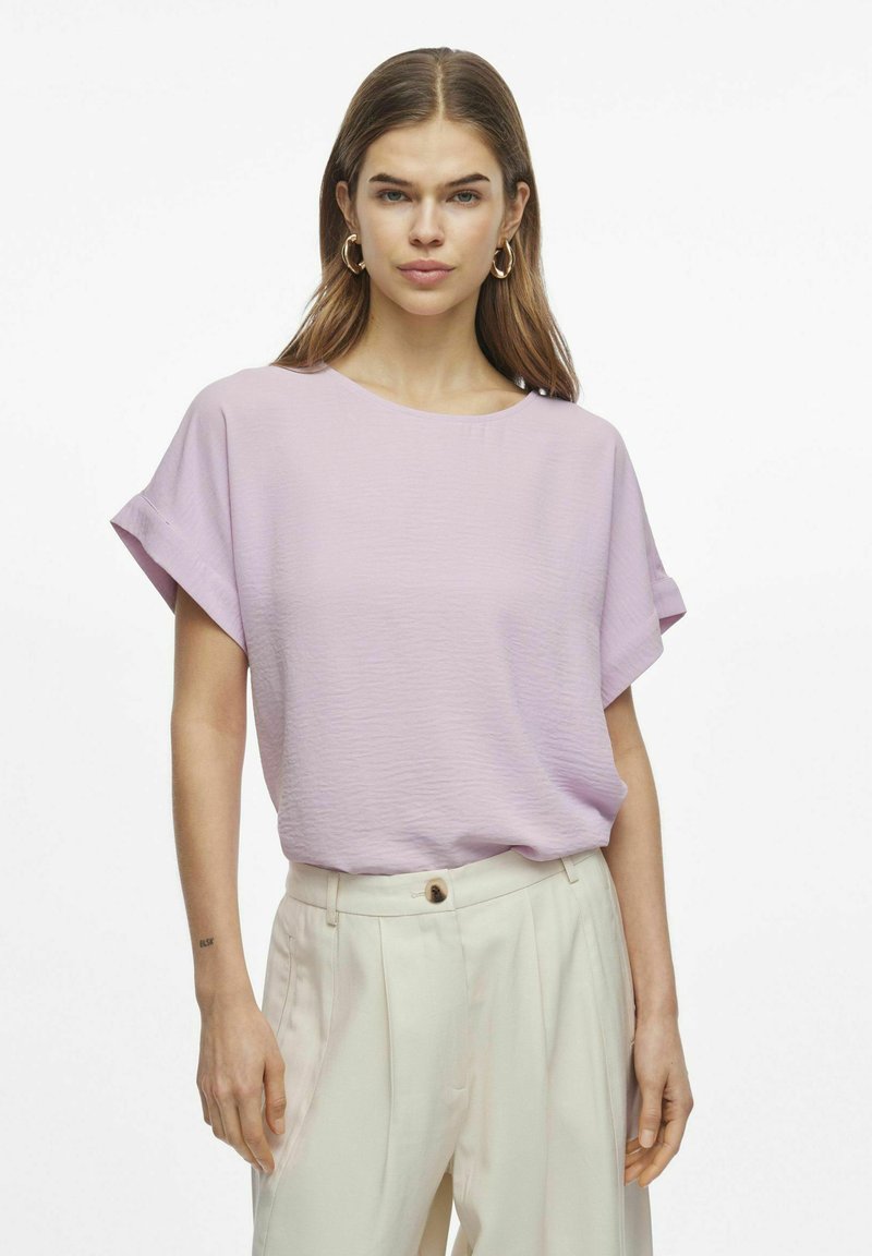 Gaiši purpura īsām piedurknēm blūze ar apaļu kaklu, kas izgatavota no teksturēta auduma. Apvienota ar gaišas krāsas augstām biksēm.